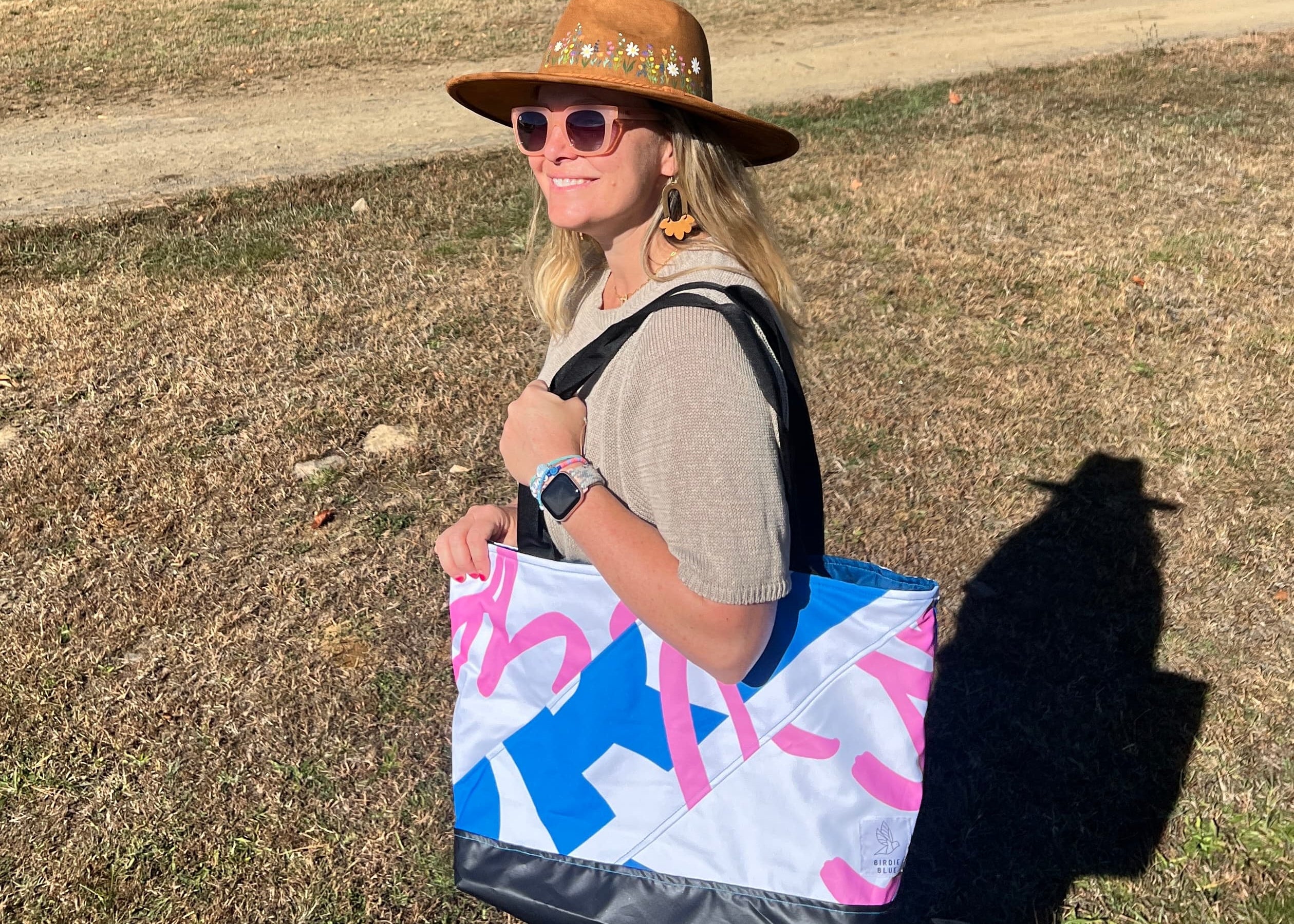Woman with a colorful tote bag and hat standing in a grassy field with trees in the background