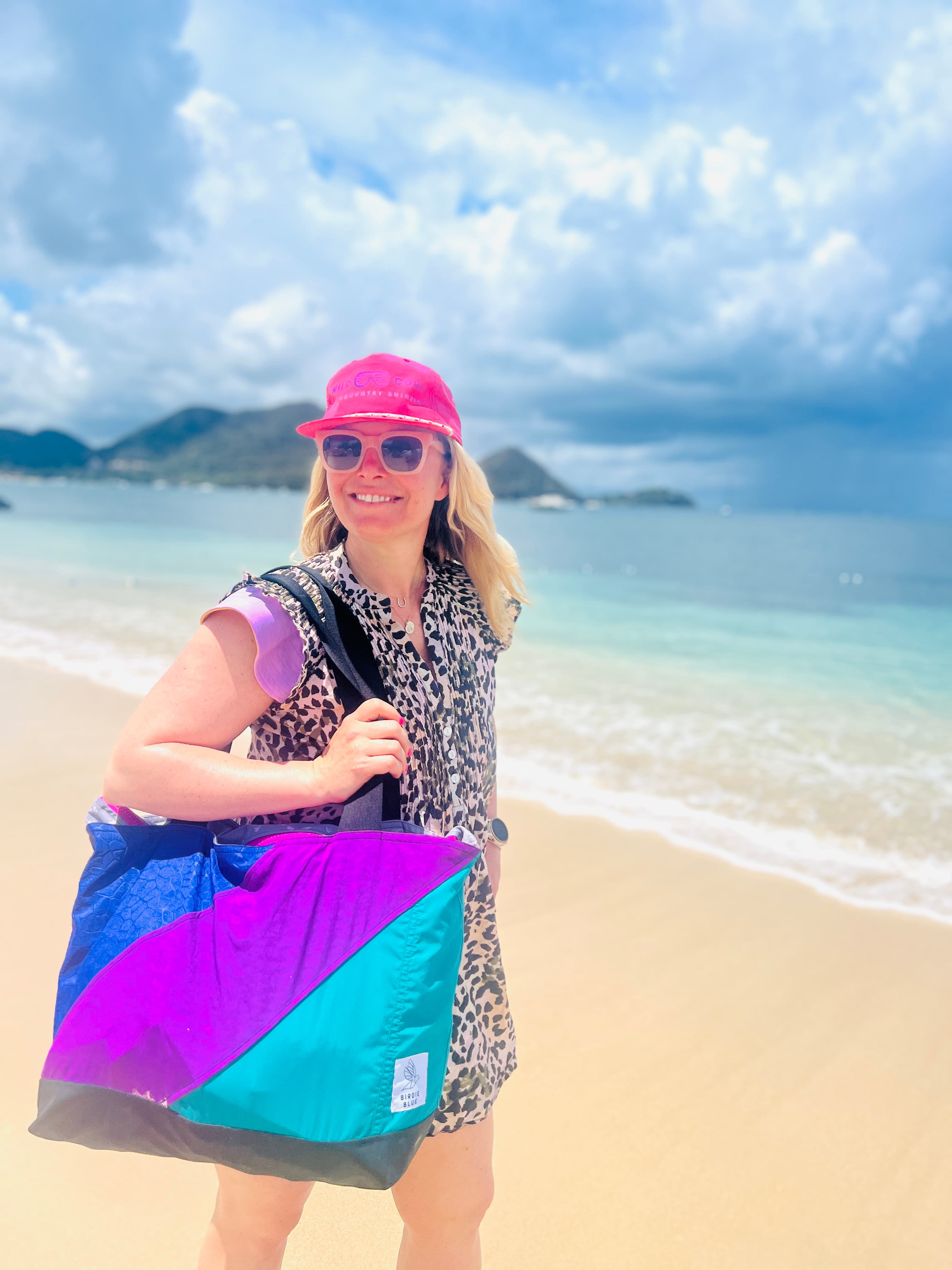 A lifestyle image of a person standing on a beach holding carry all tote, with a pink hat and sunglasses on.