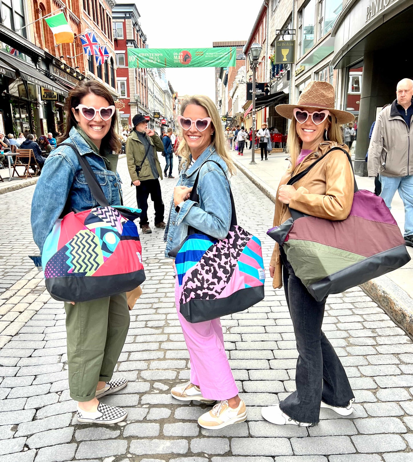 Lifestyle image of three individuals standing on a cobblestone street, each carrying a large, multicolored, Birdie Blue Carry All bag.