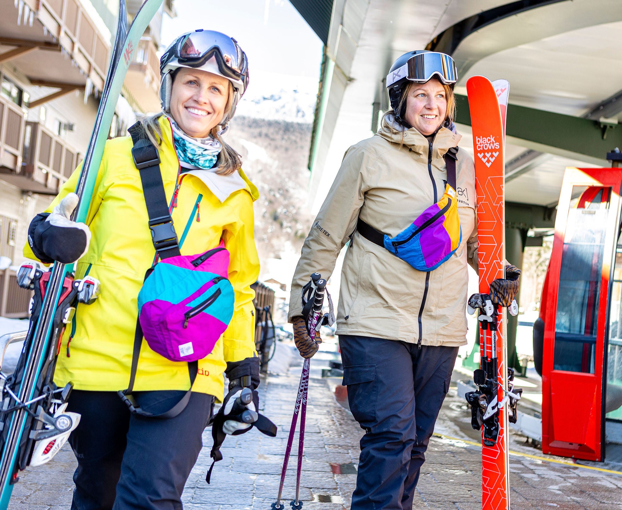 Two women carrying skis wearing colorful bags made from recycled ski gear