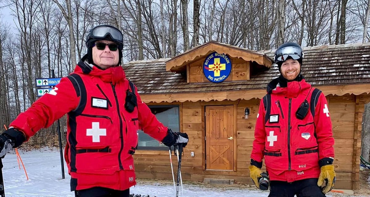 Two ski instructors in red and black Swiss Cross ski jackets standing in front of a wooden lodge in a snowy winter setting.