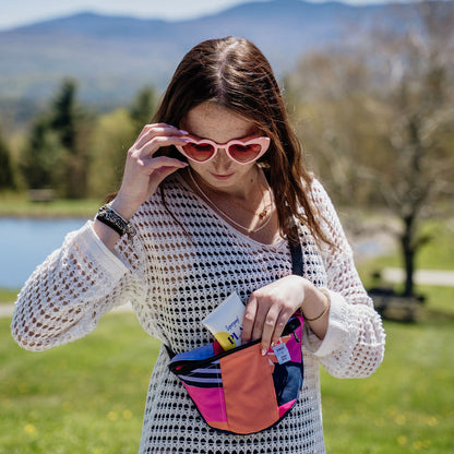 A person wearing a white mesh shirt and pink sunglasses is holding a colorful fanny pack with a red, blue, and yellow geometric pattern.