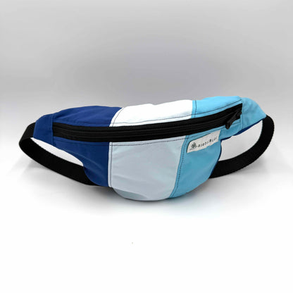 A color-blocked waist bag with panels of royal blue, white, and light blue, featuring a black adjustable strap and black zipper trim. A small white brand label is visible on the front.