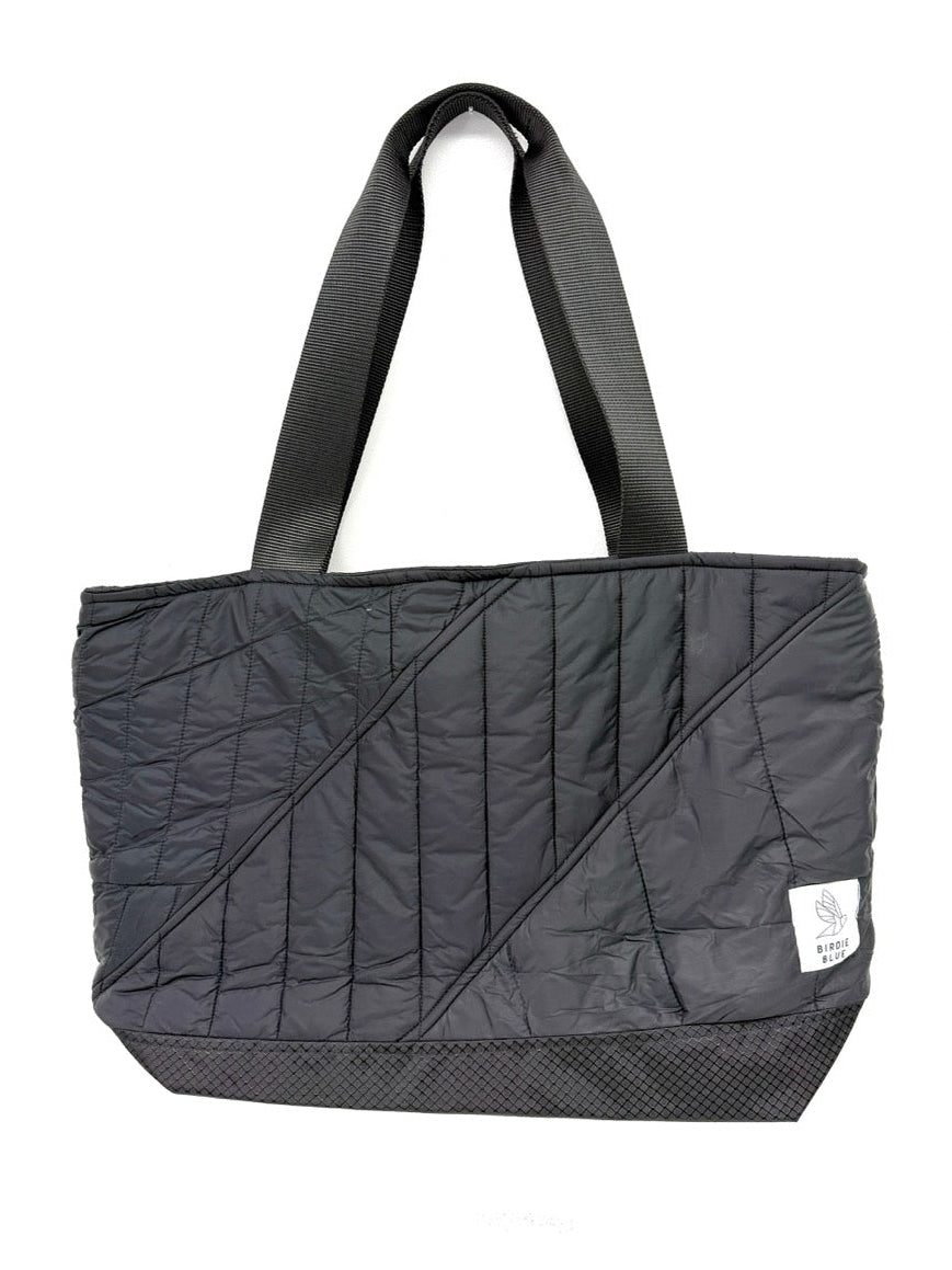 A black quilted tote bag with diagonal stitched paneling, dual shoulder straps, and a small white brand tag on the lower right side, shown against a plain white background.