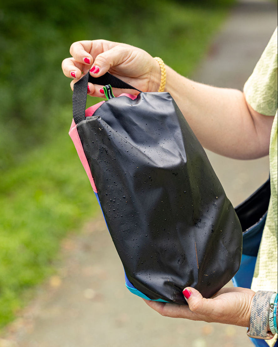 A close-up of a person outdoors holding a black waterproof pouch with pink and blue accents. The pouch has water droplets on it, and the background shows a blurred green garden path.