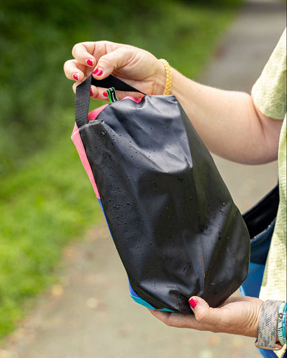 A close-up of a person outdoors holding a black waterproof pouch with pink and blue accents. The pouch has water droplets on it, and the background shows a blurred green garden path.
