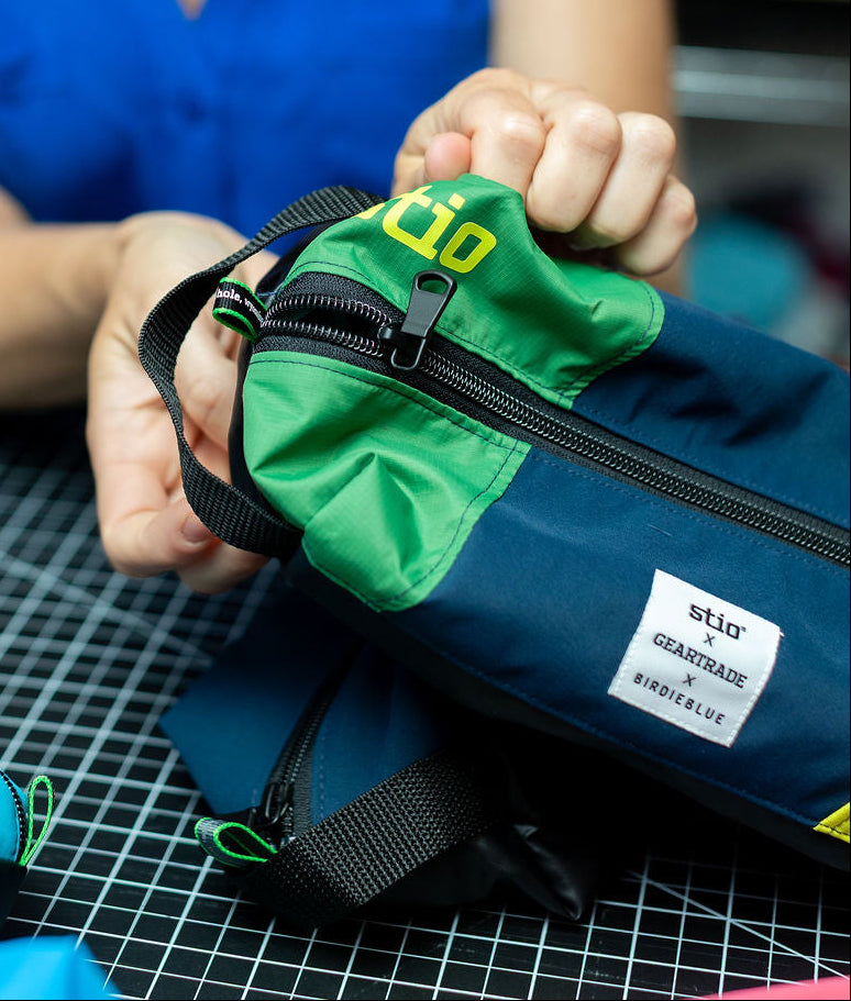 A woman in a blue sleeveless top holds a navy and green zippered pouch with a white sewn label that reads Stio x Geartrade x Birdieblue