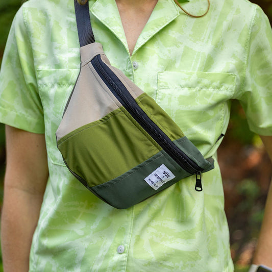 A person wearing a light green patterned short-sleeve shirt and dark pants carries a color-blocked fanny pack with olive green, beige, and dark gray panels and a black strap.